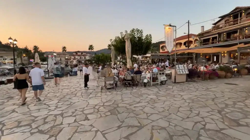 Crowded restaurant terrace spilling onto the wide stone-paved square in Sivota village at dusk, couples walking past as diners fill tables beneath a multi-storey taverna building