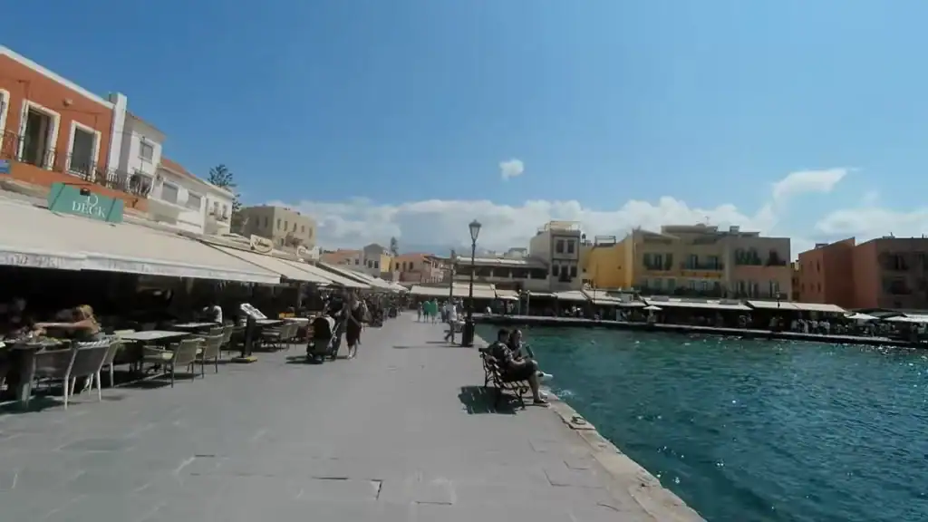 People walking and sitting along a wide stone promenade edging a harbour, restaurants with cream awnings stretching into the distance beside clear blue water