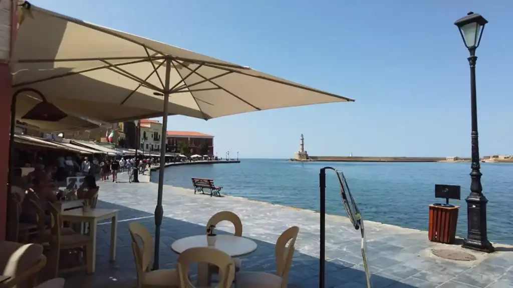 Outdoor cafe tables beneath a large cream umbrella along the waterfront promenade, with the Egyptian lighthouse visible across the harbour mouth