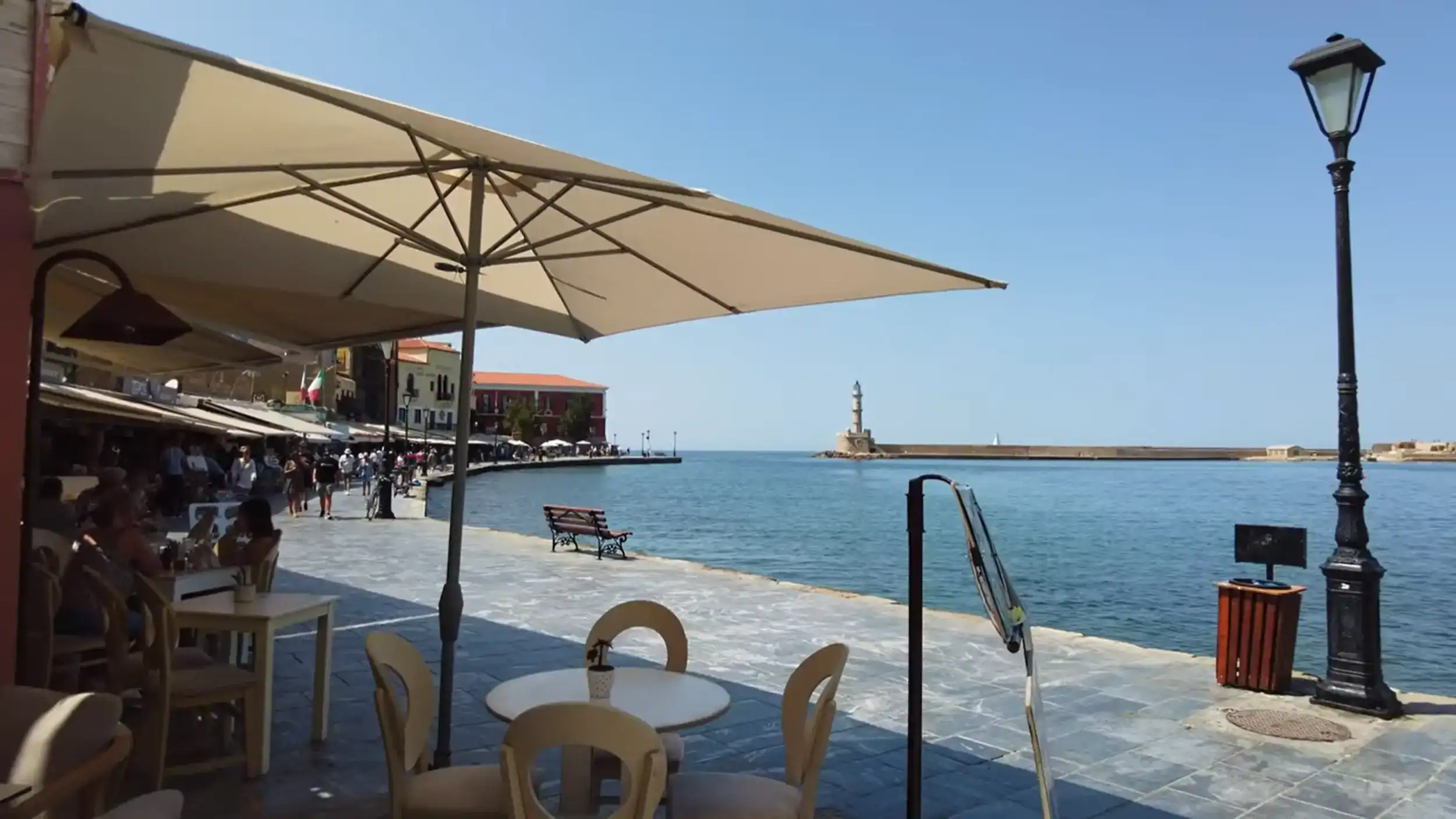 Outdoor cafe tables beneath a large cream umbrella along the waterfront promenade, with the Egyptian lighthouse visible across the harbour mouth