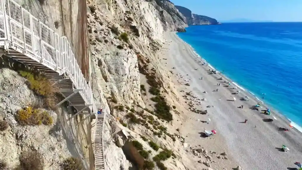 Metal staircase bolted to sheer white cliffs descending to Egremni beach, with turquoise water stretching along the shoreline below