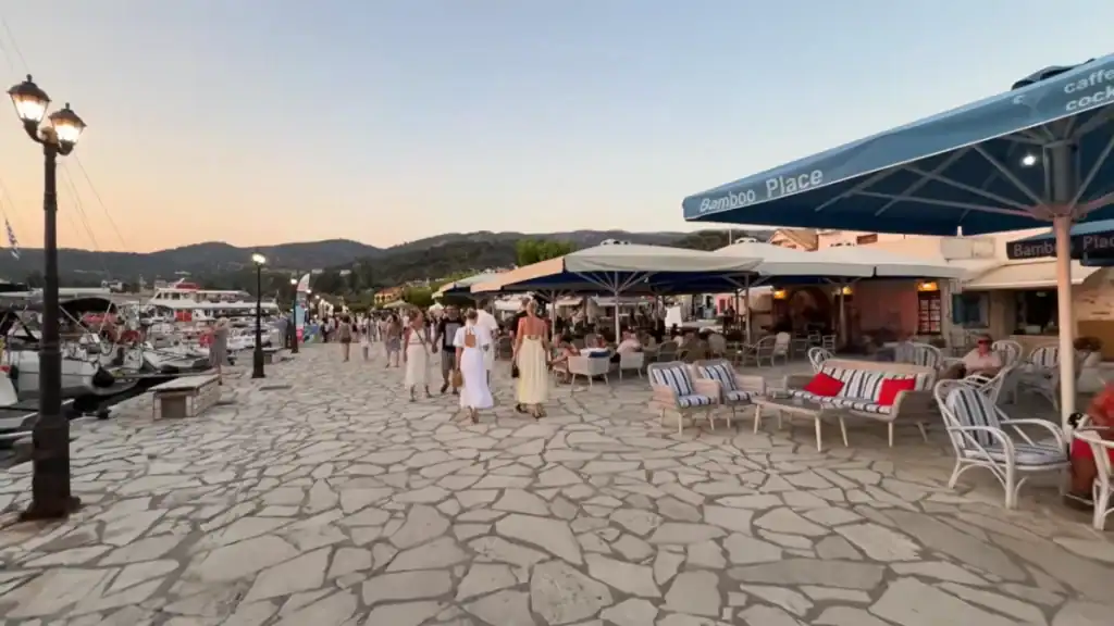 People strolling along the stone-paved harbour front in Sivota Lefkada at dusk, the Bamboo Place cafe bar terrace to the right and yachts moored along the quay to the left