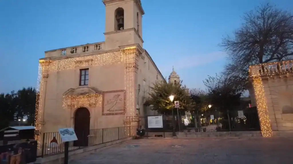 Warm fairy lights strung across the facade of the Church of Saint Vincent Ferrer in Ragusa at dusk, a sundial mounted beside the doorway and the dome of a larger church visible behind the trees.