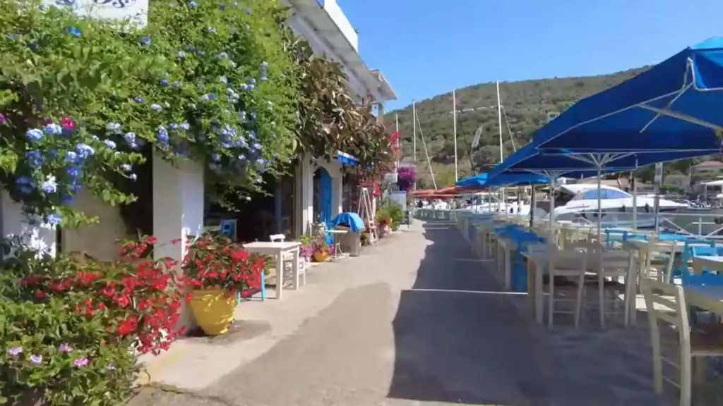 Flower-covered taverna fronts line a harbourside promenade in lefkada in may, with empty dining tables and blue parasols beside moored sailing boats