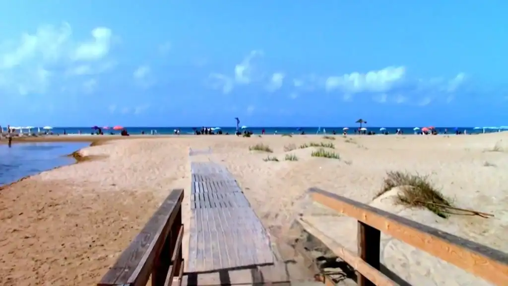 Wooden boardwalk access ramp leads onto a flat sandy beach at Marina di Modica, coloured parasols and bathers spread along the shoreline under a partly cloudy sky