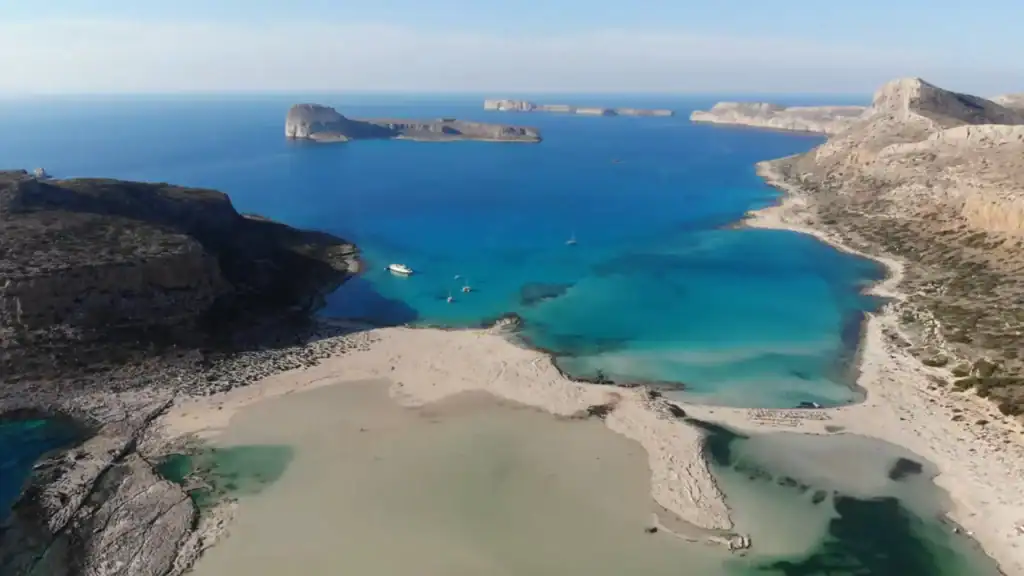 Turquoise and deep blue waters curve between pale sandy spits and rugged limestone cliffs, with small boats anchored in the shallows