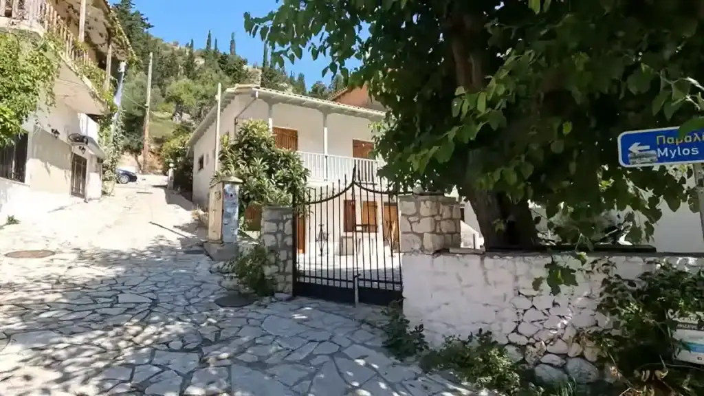 A blue directional sign reading "Mylos" points left at a cobbled junction in Agios Nikitas, with a whitewashed house behind iron gates and tree shade falling across the stone surface.