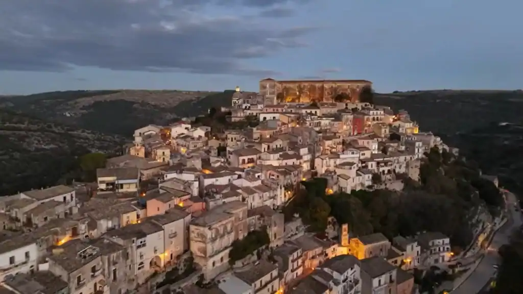 Ragusa Ibla at night, its baroque tower and dome catching the last warm glow of dusk against a fading orange sky.