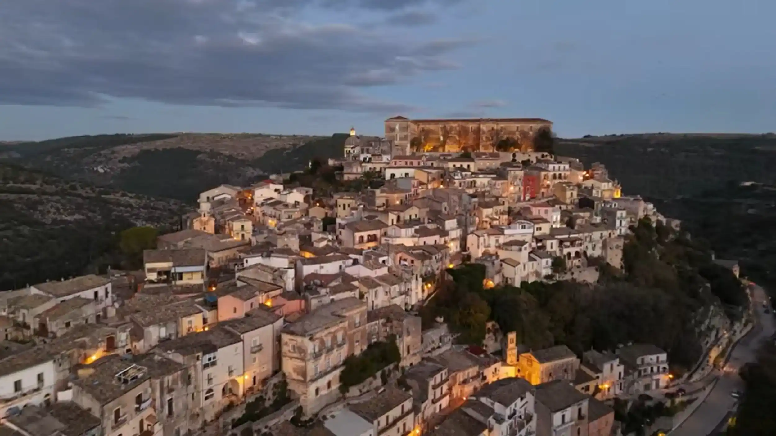 Ragusa Ibla at night, its baroque tower and dome catching the last warm glow of dusk against a fading orange sky.