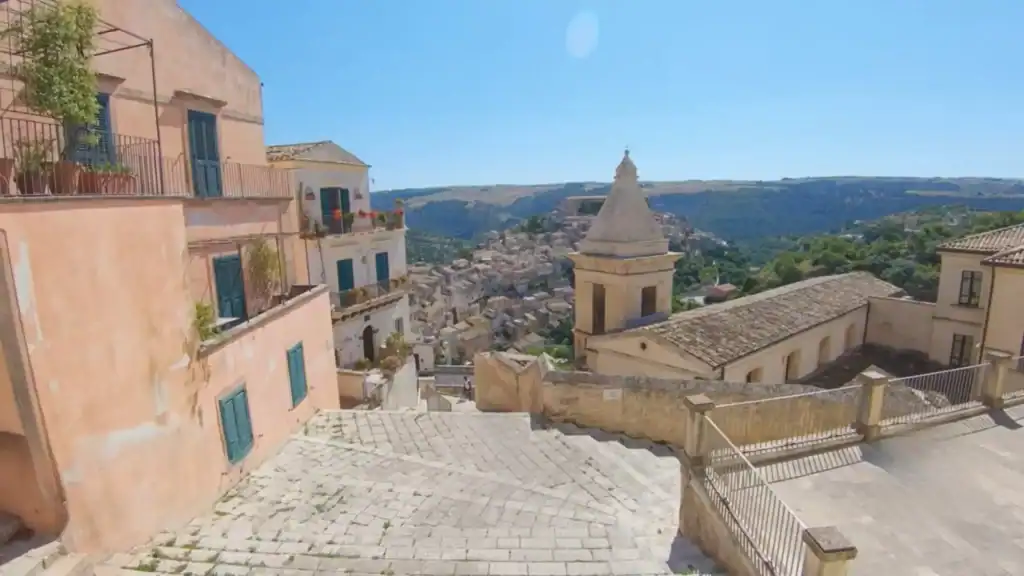 Stone steps descending past a terracotta-washed residential building toward the rooftops of Ragusa Ibla, a church cupola and the plateau escarpment beyond