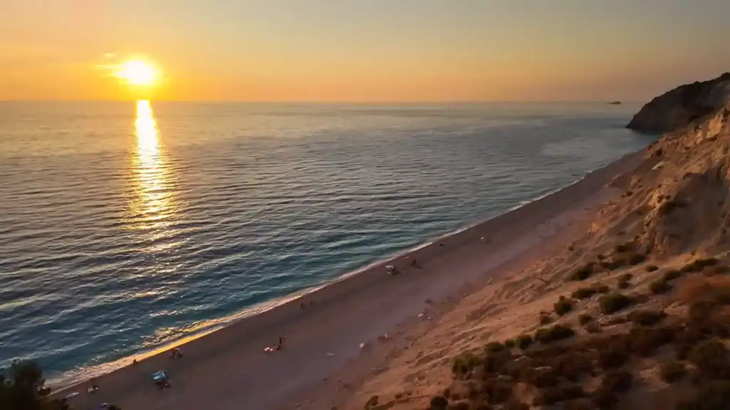 Sun setting over the Ionian Sea at Egremni, its reflection stretching across calm water on Lefkada island toward a sandy shore where a few beachgoers remain at the cliff's base.