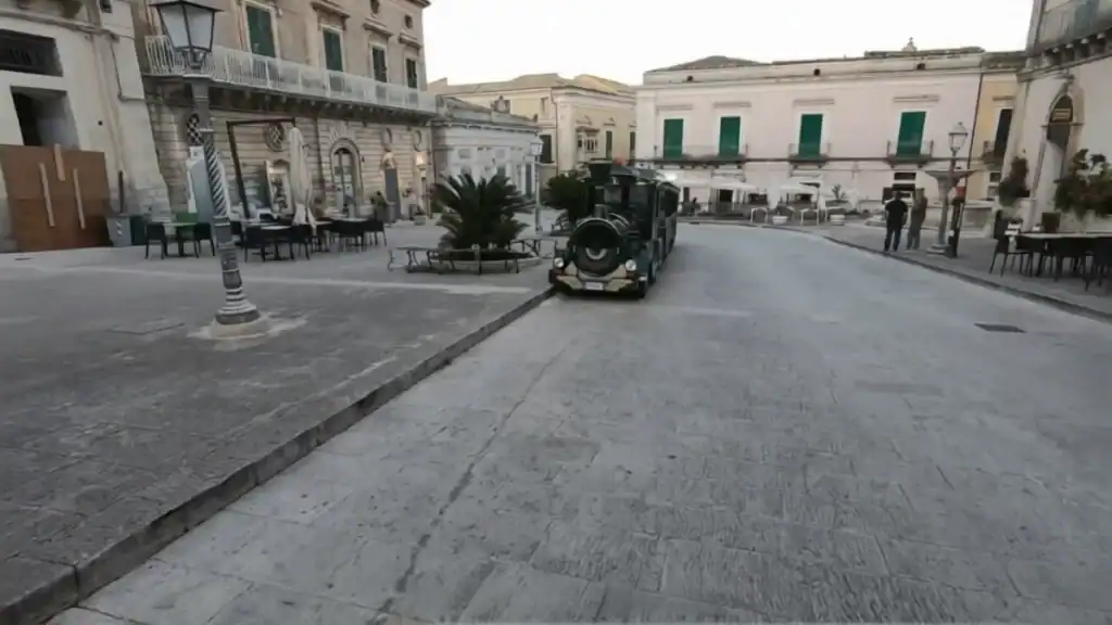 Small tourist road train passing through an almost empty piazza in Ragusa Ibla, baroque buildings and a cycad palm behind it