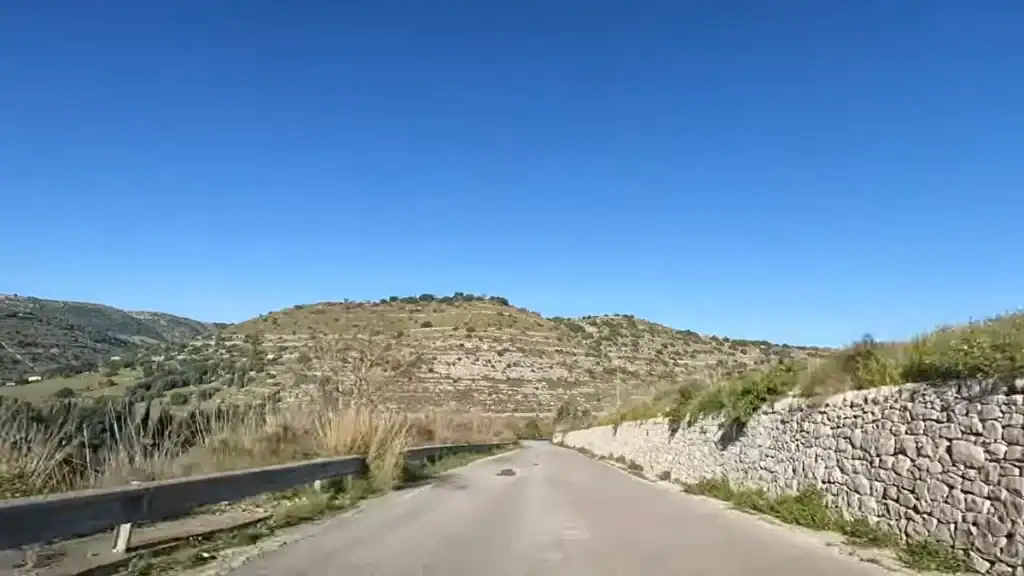 Two-lane road running toward a bare limestone hill, dry scrub to the left and a roughly coursed stone wall to the right.