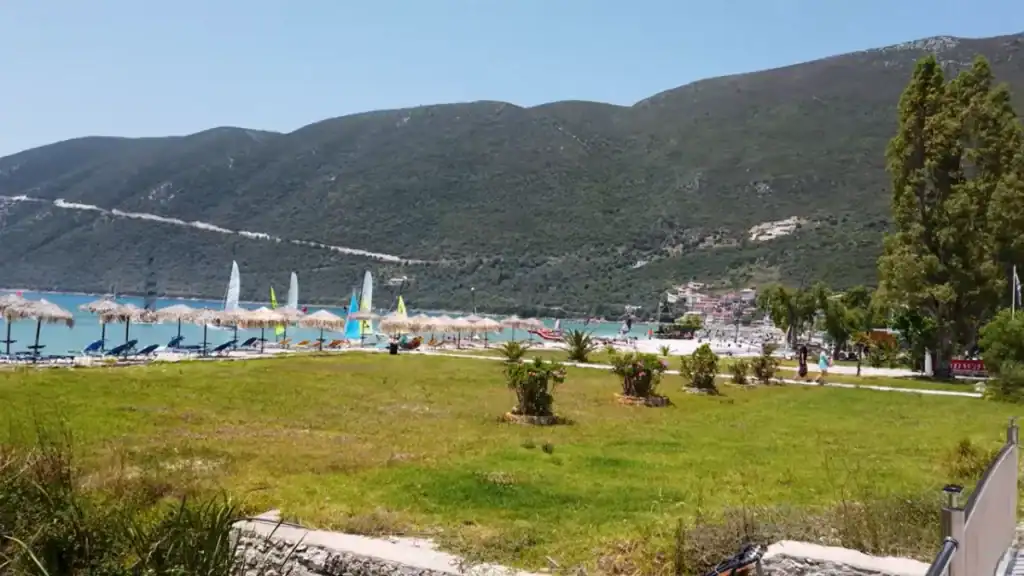 Vassiliki bay seen from a grassed area, thatched umbrellas and windsurfing sails lined along the shore with the village and green hills behind