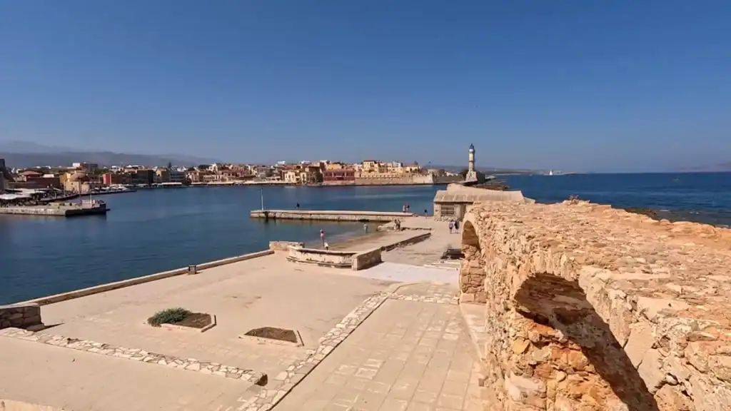 Chania old harbour seen from the Firkas fortress walls, the Egyptian lighthouse standing at the breakwater tip with the old town's coloured facades stretching behind