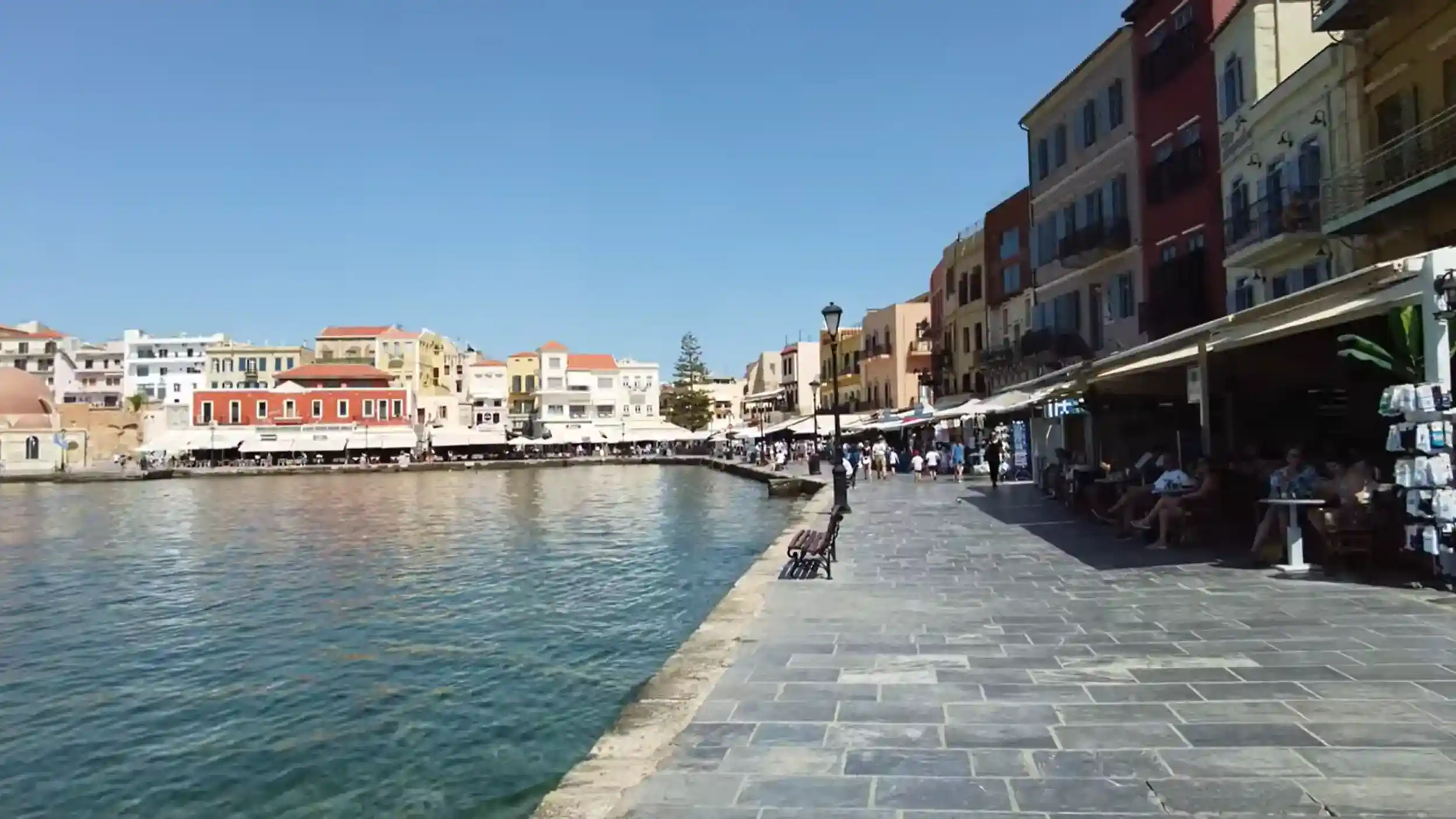 Aerial view of a curved harbour with a stone lighthouse at the end of a breakwater, colourful waterfront buildings encircling turquoise water