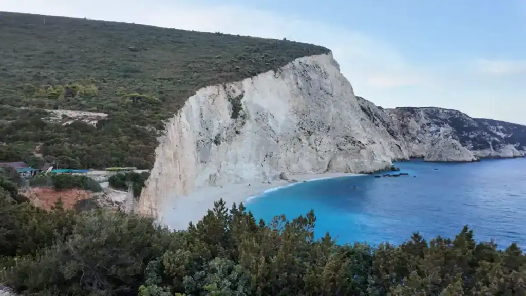 Pale limestone cliff dropping to a narrow sand beach with vivid turquoise water, forested slopes rising behind and a small structure visible at the base.