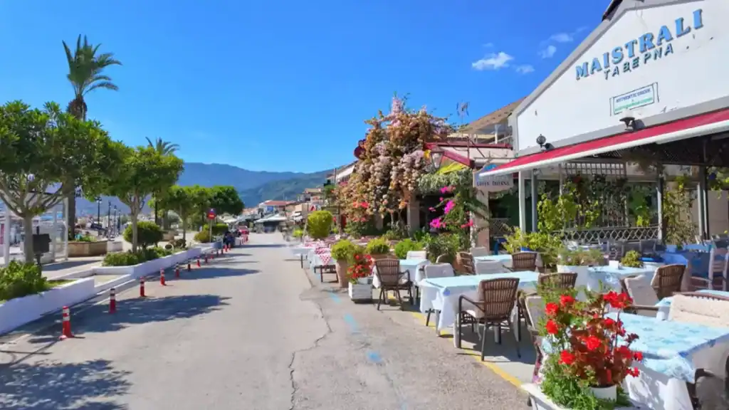 Maistrali taverna's flower-covered exterior and empty outdoor tables set with blue cloths along the main Nidri Lefkada waterfront road, palm trees lining the opposite pavement and hills stretching into the distance