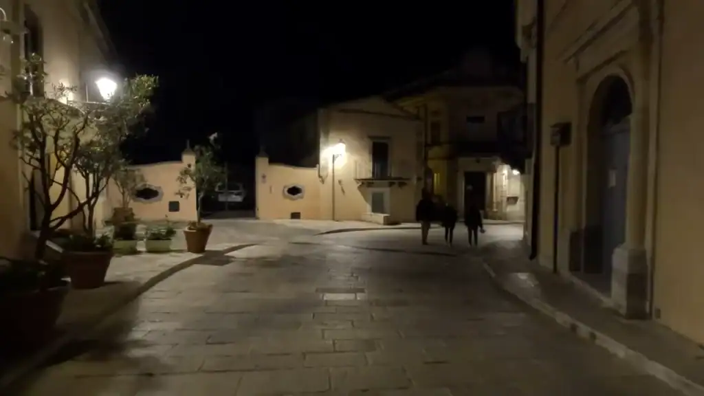 Three figures walking a curved cobbled lane in Ragusa Ibla at night, warm-walled buildings and potted trees catching the lamplight