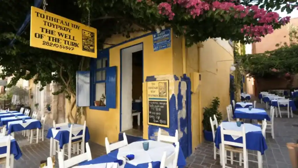 The Well of the Turk restaurant in Chania's old town, its yellow facade and blue shutters framed by bougainvillea, tables with blue cloths set across the cobbled courtyard - a find that rewards solo travel in Chania.