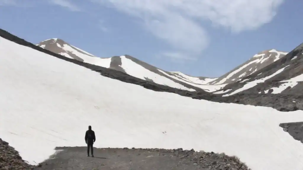 Solo hiker standing on a rocky path beside vast snow-covered slopes, multiple peaks rising against a partly cloudy sky