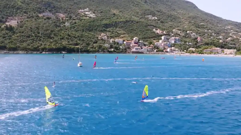 Windsurfers with brightly coloured sails cutting across the bay in Vasiliki Lefkada, the village and its tree-covered hillside stretching along the far shore.