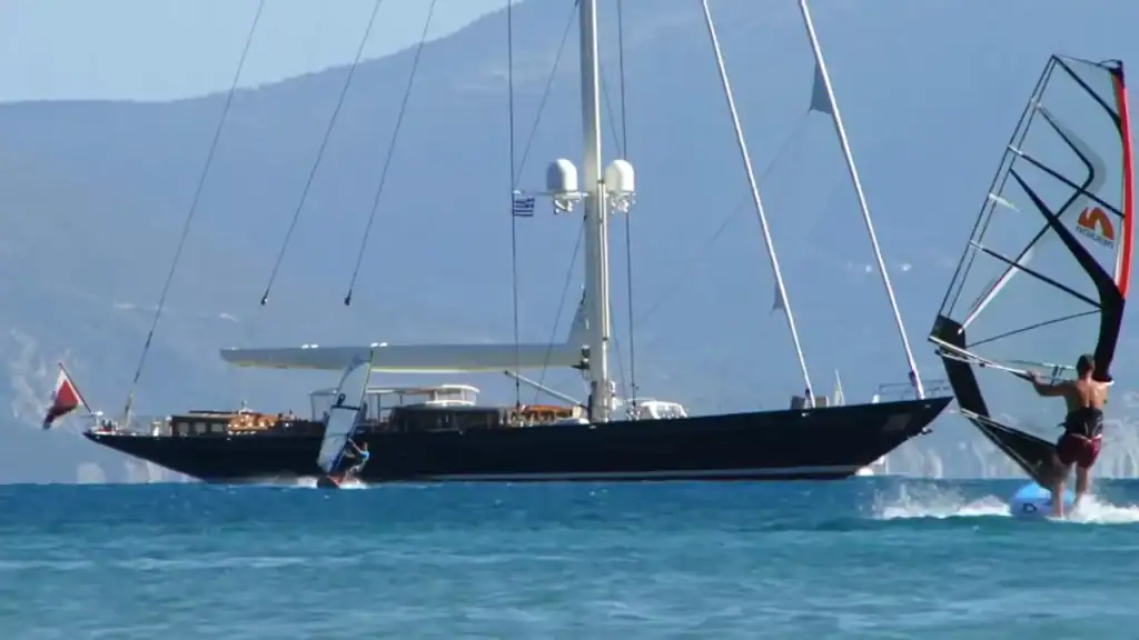 Windsurfer cutting across choppy water beside a large dark-hulled sailing yacht flying a Greek flag, with mountain slopes and white cliffs receding into haze across the bay.