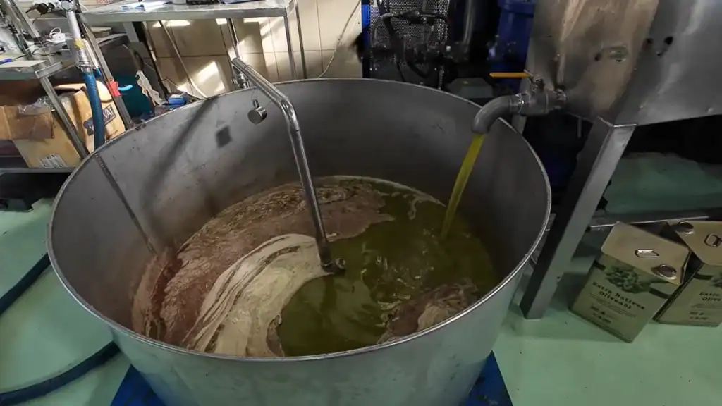 Bright green-gold olive oil pouring from a metal tap into a large steel vat filled with swirling oil and olive paste, inside a working press facility