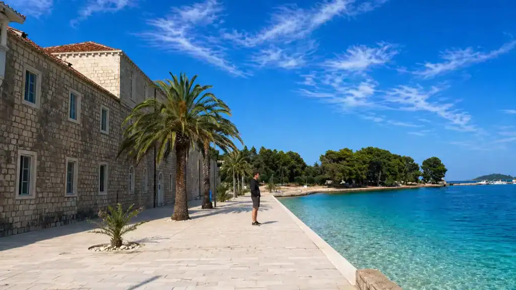 Man standing on the waterfront terrace of Badija Island monastery beside palm trees and turquoise water