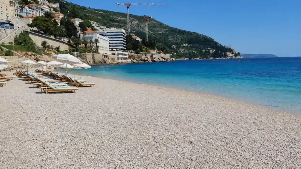 Rows of wooden sunloungers and white parasols on the fine pebble shore at Banje Beach with a hotel and hillside behind