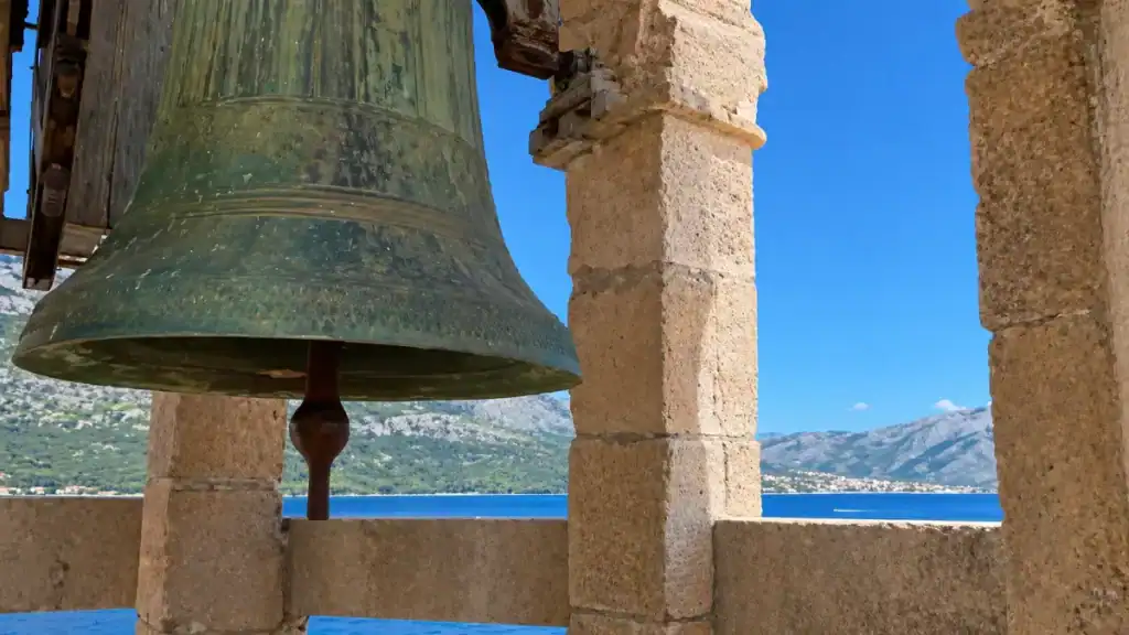 Large bronze bell atop a stone tower with the Pelješac Peninsula mountains and Adriatic Sea beyond