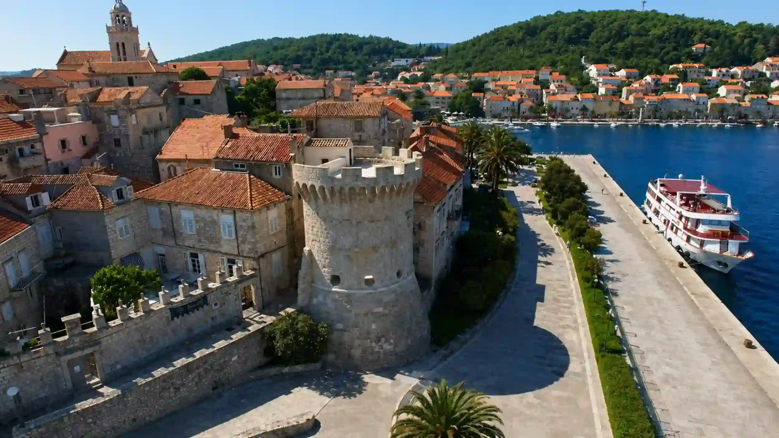 Aerial view of Korčula town's medieval tower, old town rooftops, and harbour with a cruise ship docked alongside