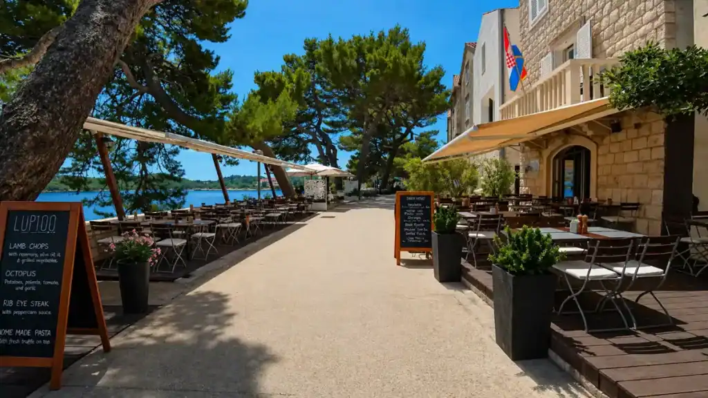 Waterfront restaurant terrace with outdoor tables, pine trees, and sea views along a shaded promenade