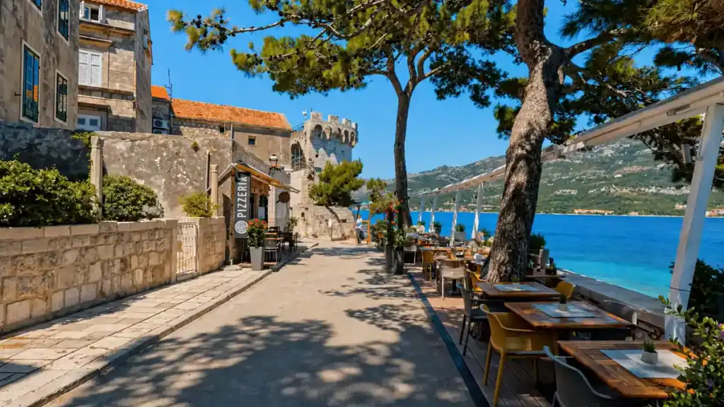 Waterfront path in Korčula Town with outdoor restaurant tables and a medieval tower beyond