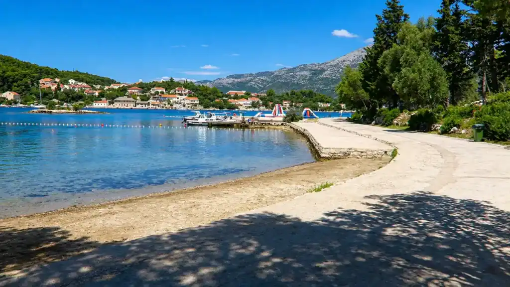 Shaded promenade along Korčula's eastern bay with moored boats, inflatable water park, and hillside village across the water