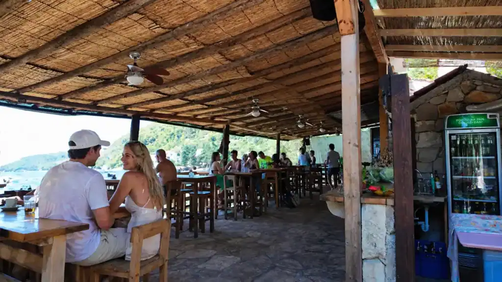Diners under a reed-roof beach bar at Pupnatska Luka with views across the cove to pine-covered hills