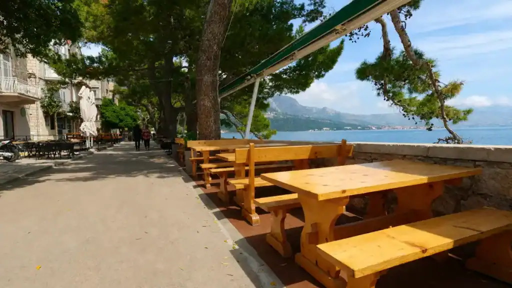 Empty wooden benches at a seafront bar terrace on Korčula's promenade, Pelješac peninsula mountains visible across the channel