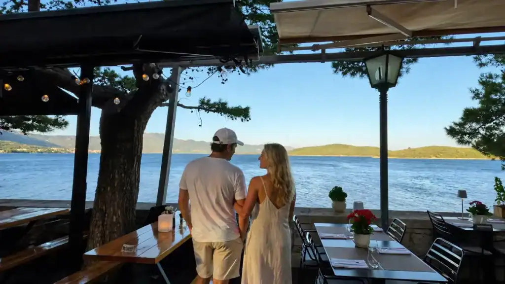 Couple standing at silk restaurant terrace in Korcula overlooking calm sea and a green island at dusk.