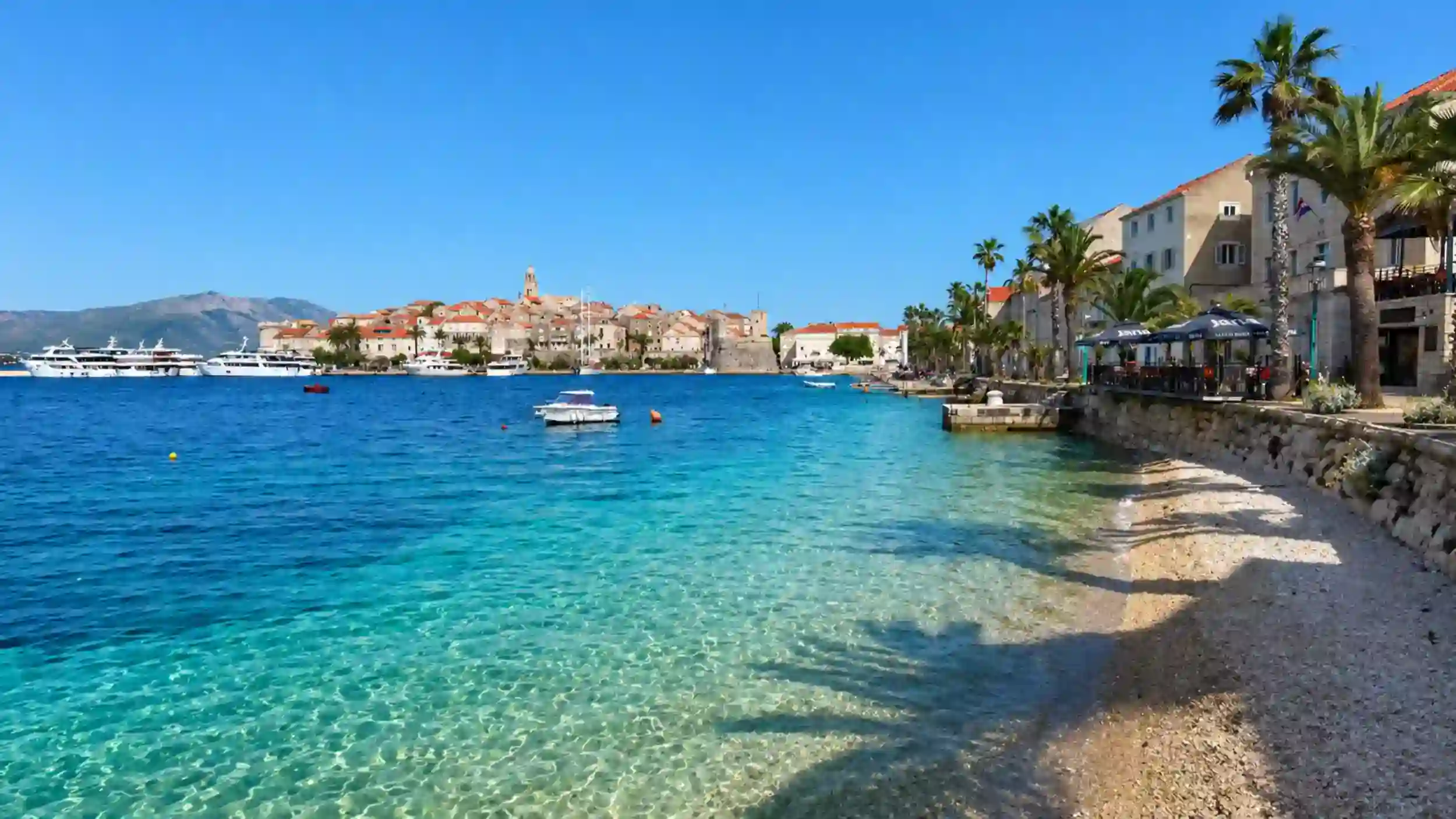 Shallow pebble shore on Korčula's northern bay with the walled old town skyline, cathedral tower, and moored yachts across the water