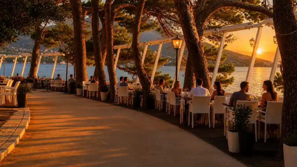 Diners seated at a waterfront restaurant terrace during Korčula town sunset, pine trees lining the promenade