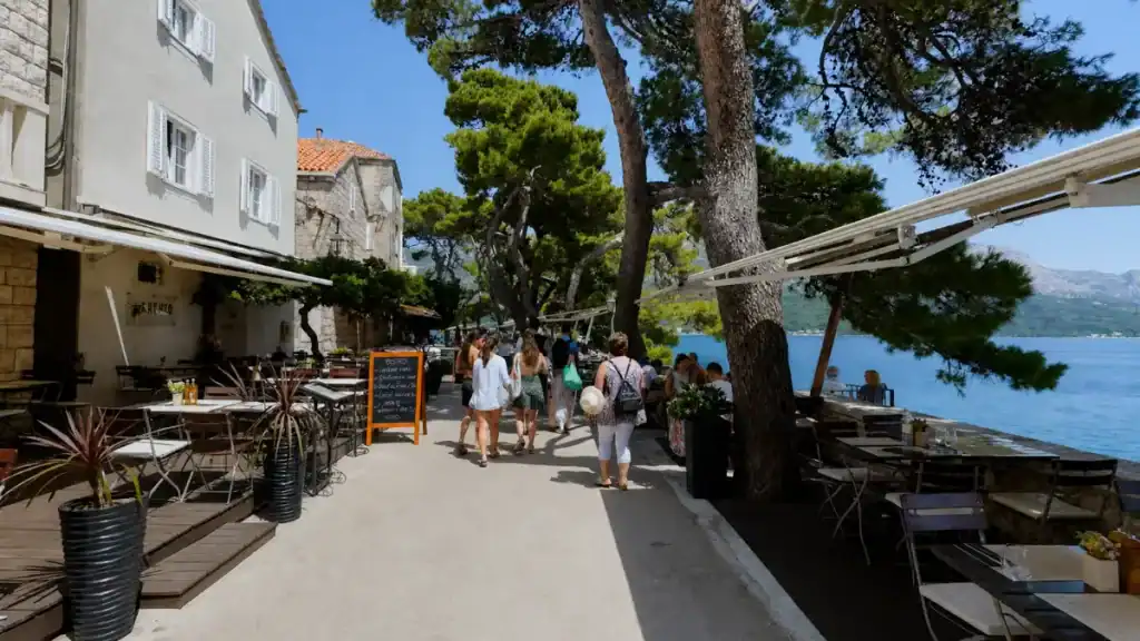 Visitors walking along the shaded waterfront promenade beside outdoor restaurant terraces in Korčula old town