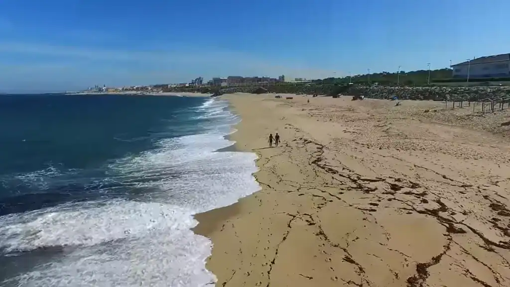Two people walking the shoreline of Vila do Conde beach with the town skyline stretching north
