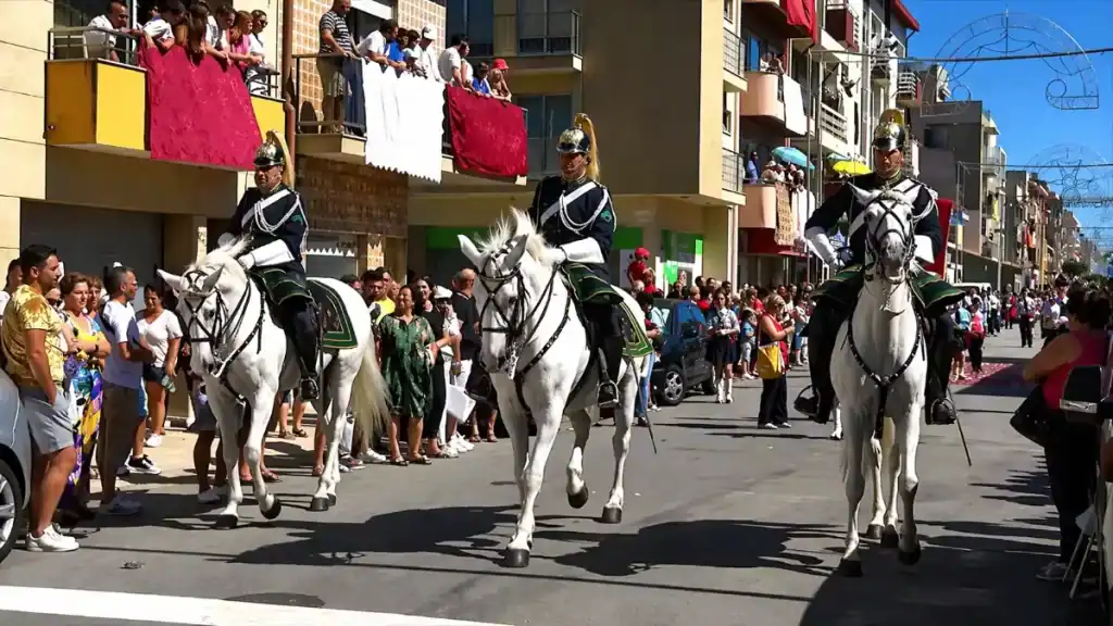 Three uniformed cavalry guards on white horses riding past crowded balconies along a town street