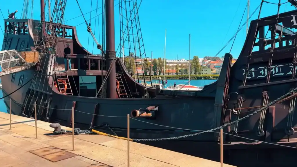 Dark-hulled replica caravel moored at a riverside quay with rigging and a cannon visible