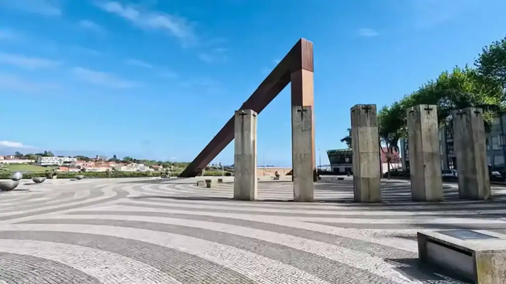 Large steel sundial gnomon and concrete pillars on a patterned cobblestone plaza by the waterfront