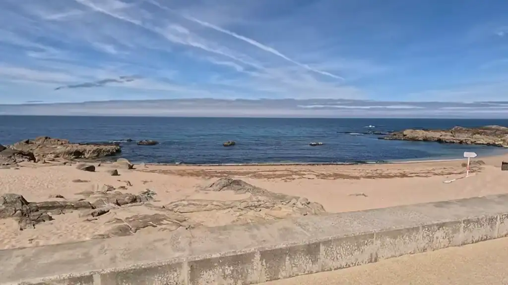 Empty sandy cove flanked by granite outcrops and a low sea wall under a pale blue sky