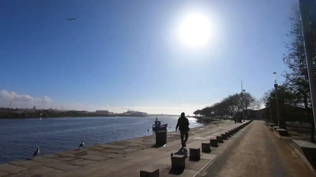 Lone figure walking along the sunny river Ave promenade with a fishing boat moored nearby.