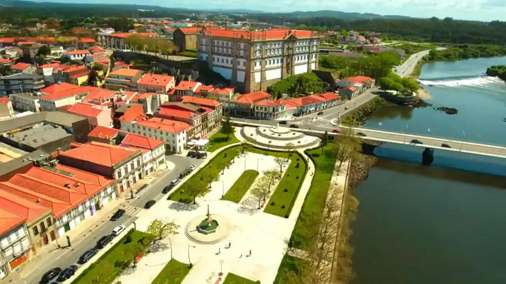 Aerial shot of a riverside town square with manicured gardens, a bridge, and terracotta rooftops