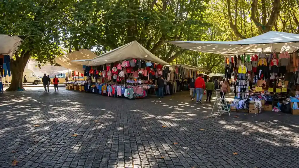 Outdoor market stalls selling colourful clothing beneath a canopy of mature plane trees on cobblestones