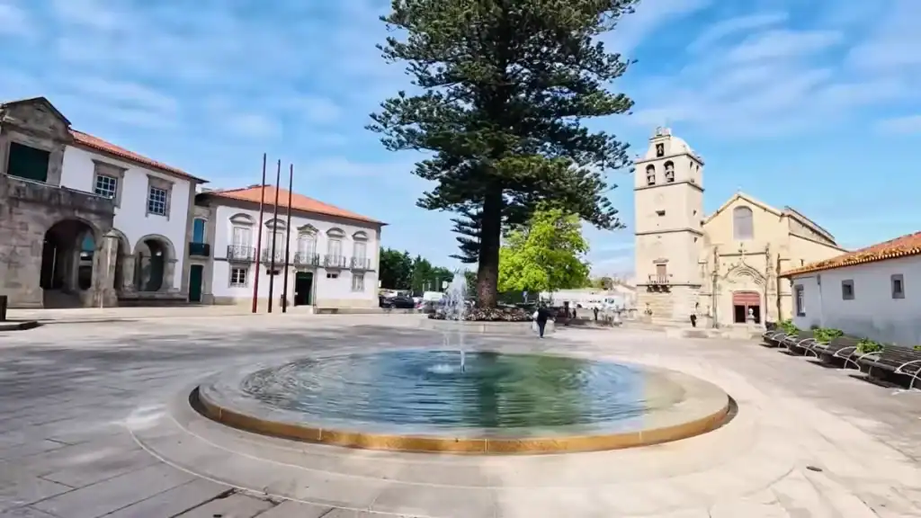 Circular fountain in an open town square flanked by a stone church tower and whitewashed civic buildings